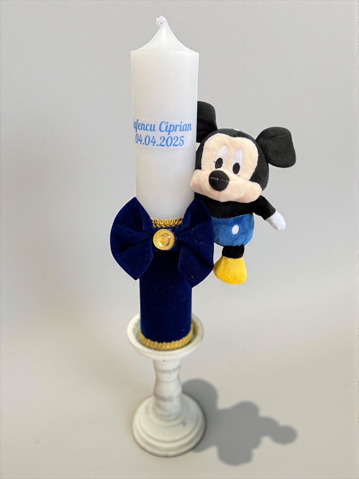 Lumânare albă personalizată cu numele 'Ciprian' și data '04.04.2025', decorată cu un fundiță albastră și o jucărie Mickey Mouse.